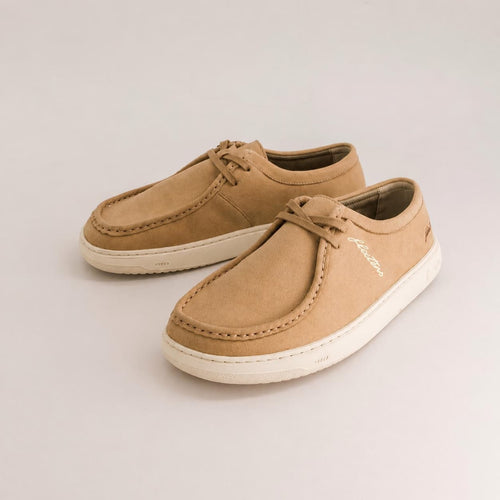 A pair of tan suede slip-on shoes with fringe details and white soles, photographed on a clean light gray background.