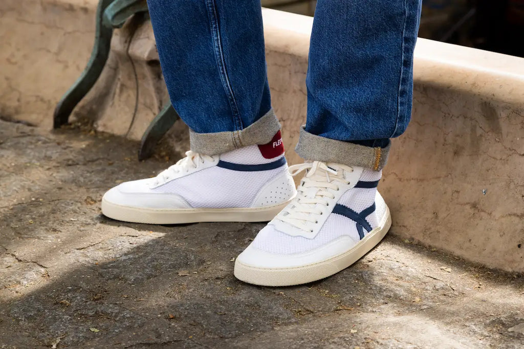 White mid-ankle barefoot sneakers with blue accents worn by a person sitting on a stone bench.