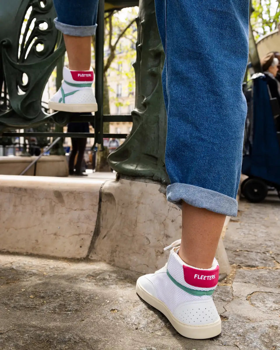 Close-up of feet wearing mid-ankle barefoot sneakers with colorful soles and green rain boots on a concrete surface.