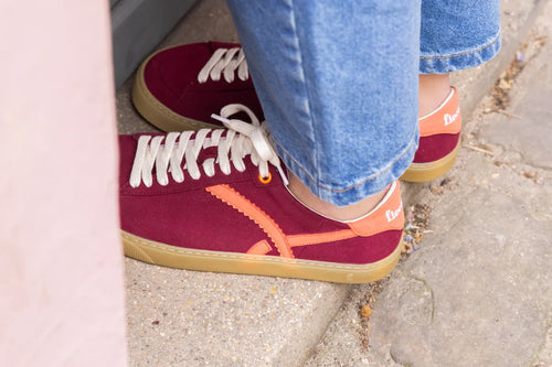 Red minimalist sneakers with orange accents worn with blue jeans on a concrete surface.