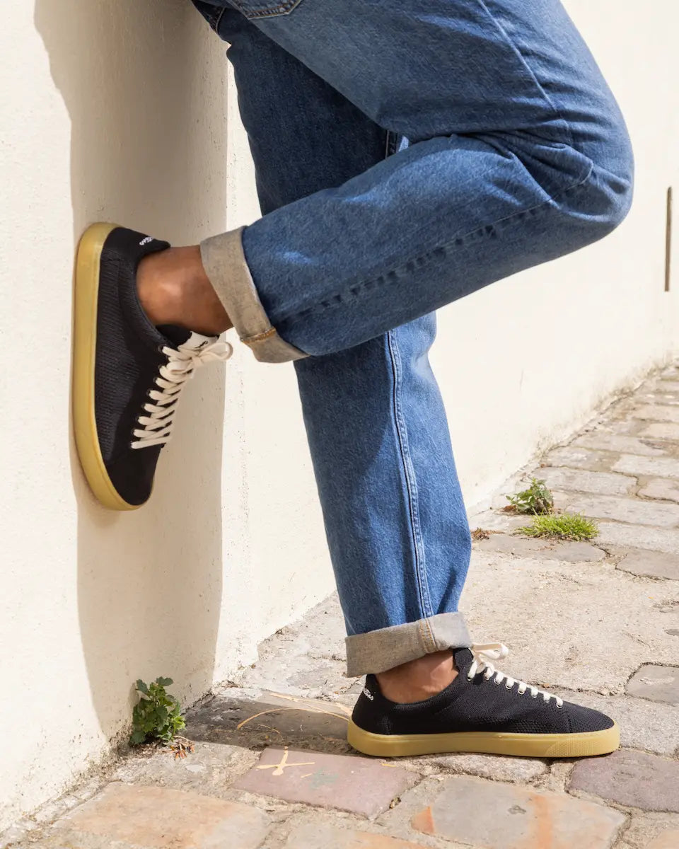 Person wearing black knit barefoot sneakers with yellow soles and blue jeans on a stone pavement.