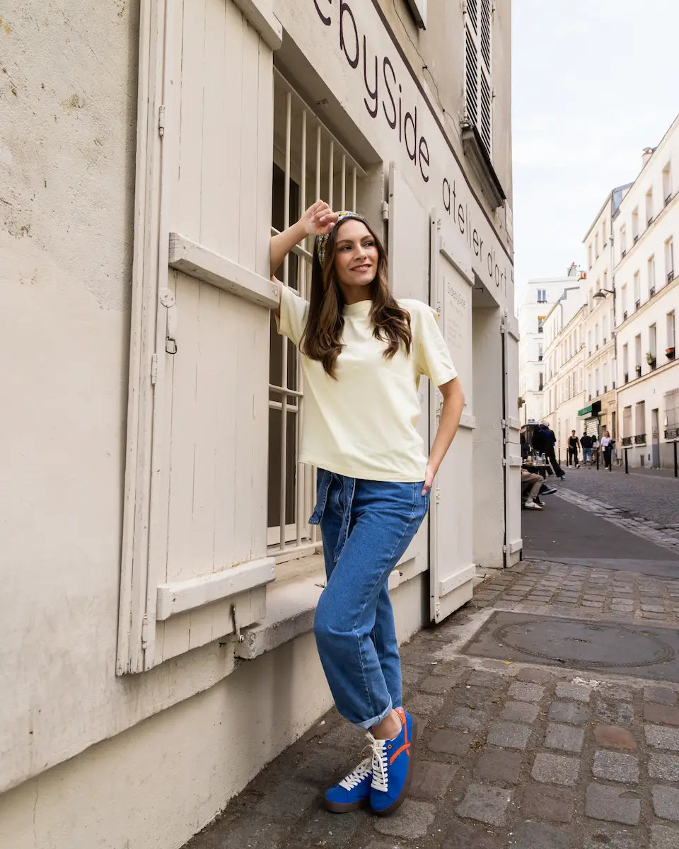 Woman standing on a street in an urban setting, wearing a light-colored top and blue jeans.