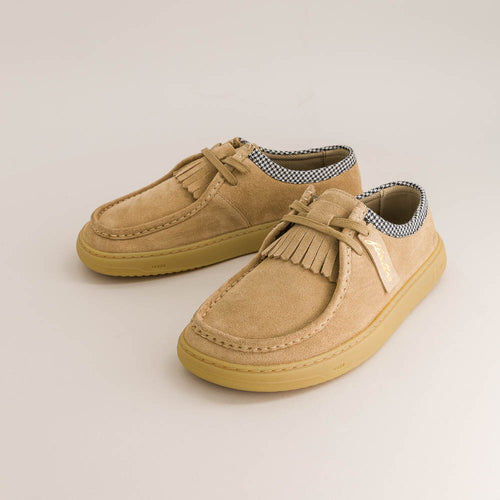 Pair of tan suede barefoot derby shoes with fringes on a light beige background