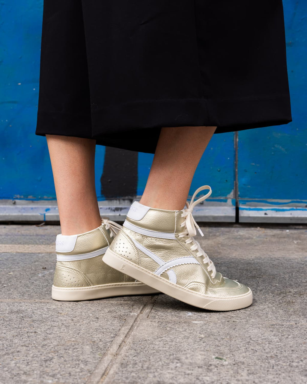 A person wearing metallic gold high-top barefoot sneakers with white side stripes and white laces, standing on a concrete sidewalk in front of a blue wall.