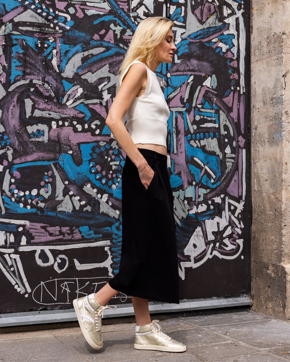 A woman stands in profile against a colorful graffiti wall, wearing a white sleeveless crop top, a long black skirt, and metallic gold barefoot sneakers.