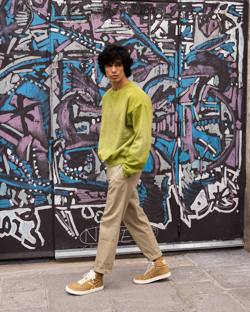 A man wearing a lime green crewneck sweatshirt, beige trousers, and tan barefoot sneakers stands in front of a graffiti-covered metal door.