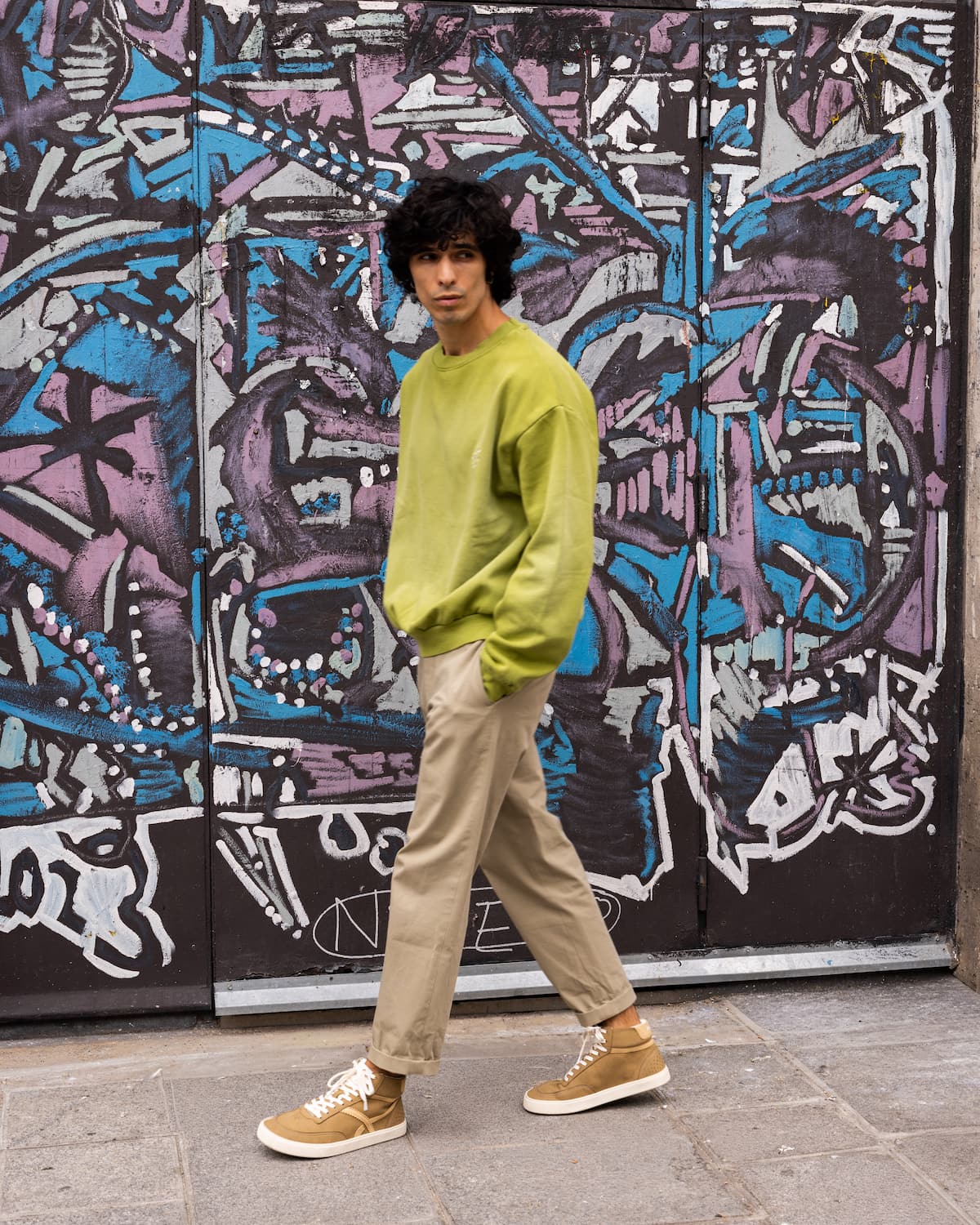 A man wearing a lime green crewneck sweatshirt, beige trousers, and tan barefoot sneakers stands in front of a graffiti-covered metal door.