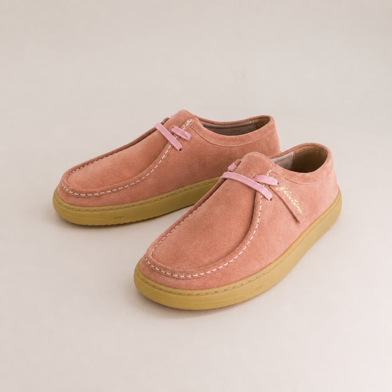 A pair of pink suede slip-on shoes with light pink laces and tan rubber soles, displayed on a light beige background.