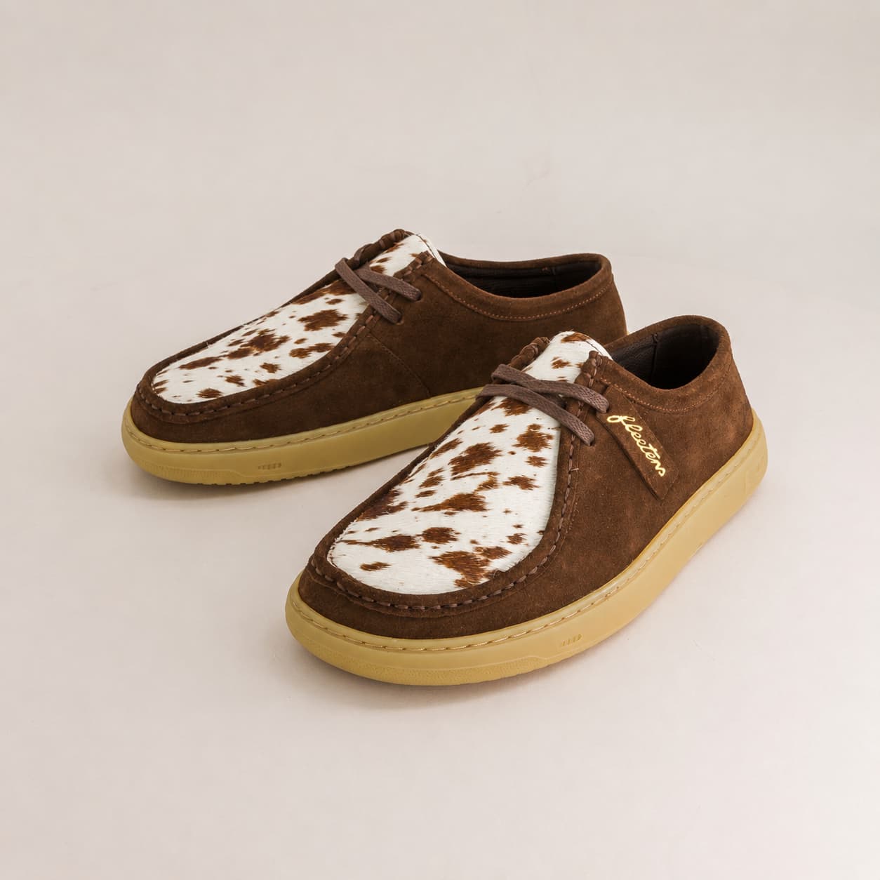 A pair of brown suede shoes with white cow-print vamps and tan soles, displayed on a light beige background.