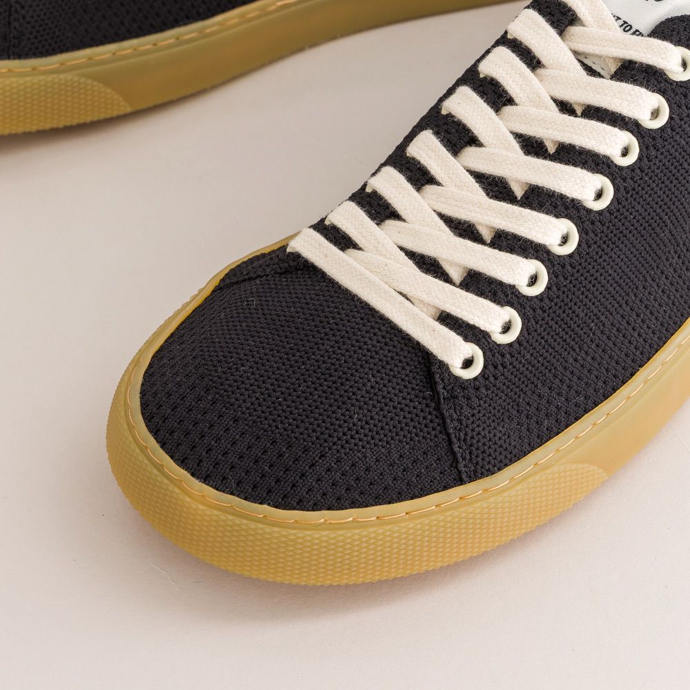 Black knit barefoot sneaker with white laces and yellow sole on a light background