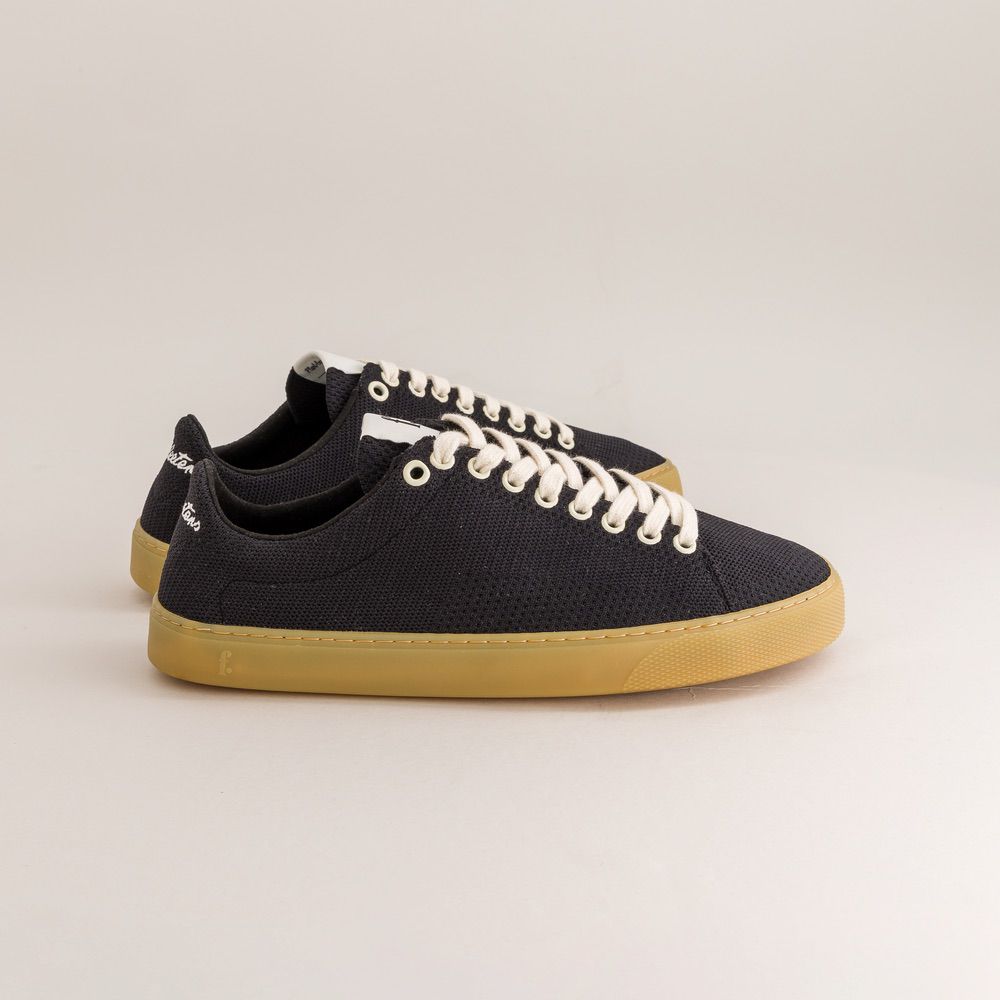 Black knit barefoot sneakers with white laces and tan soles on a light gray background