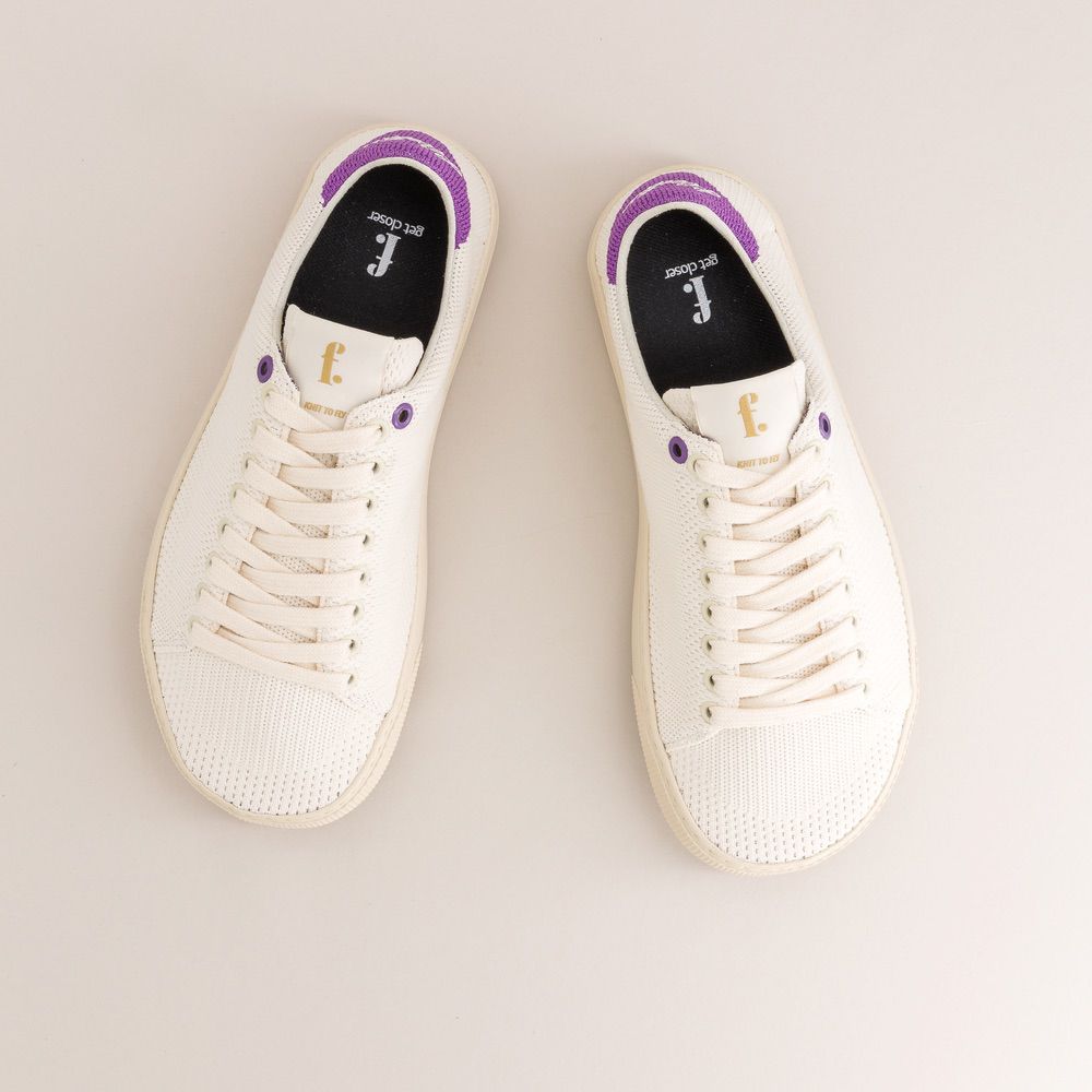 Pair of white knit barefoot sneakers with purple accents on a light gray background