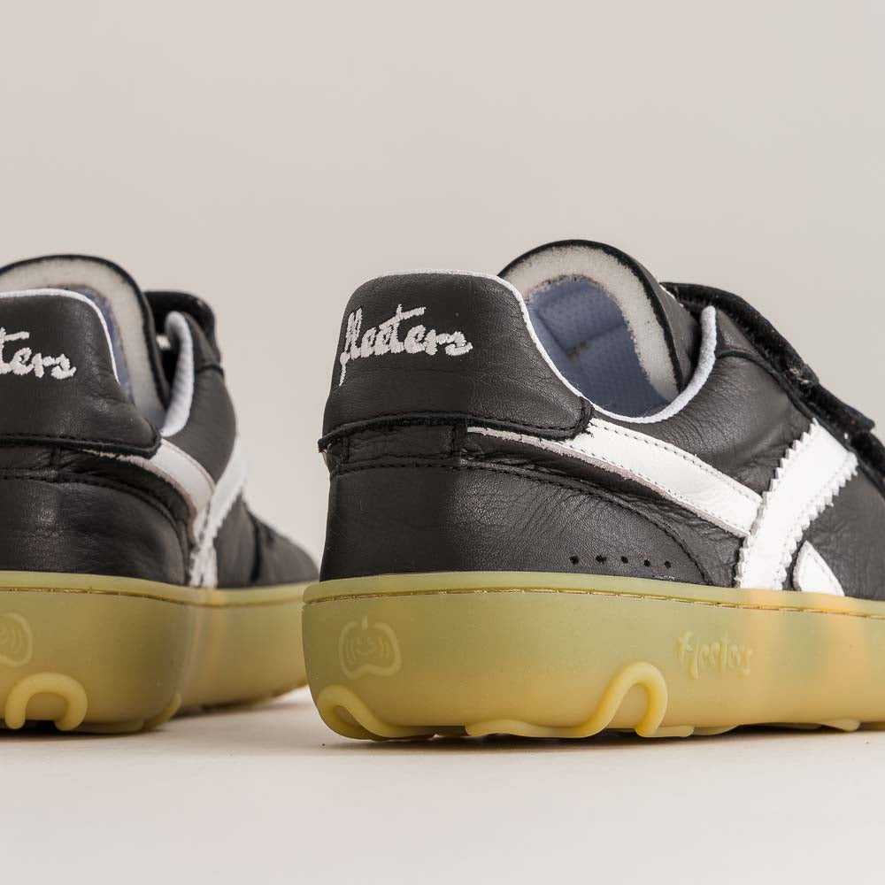 Close-up of a pair of black leather sneakers with white side stripes and translucent yellow soles, photographed on a clean light beige studio background.