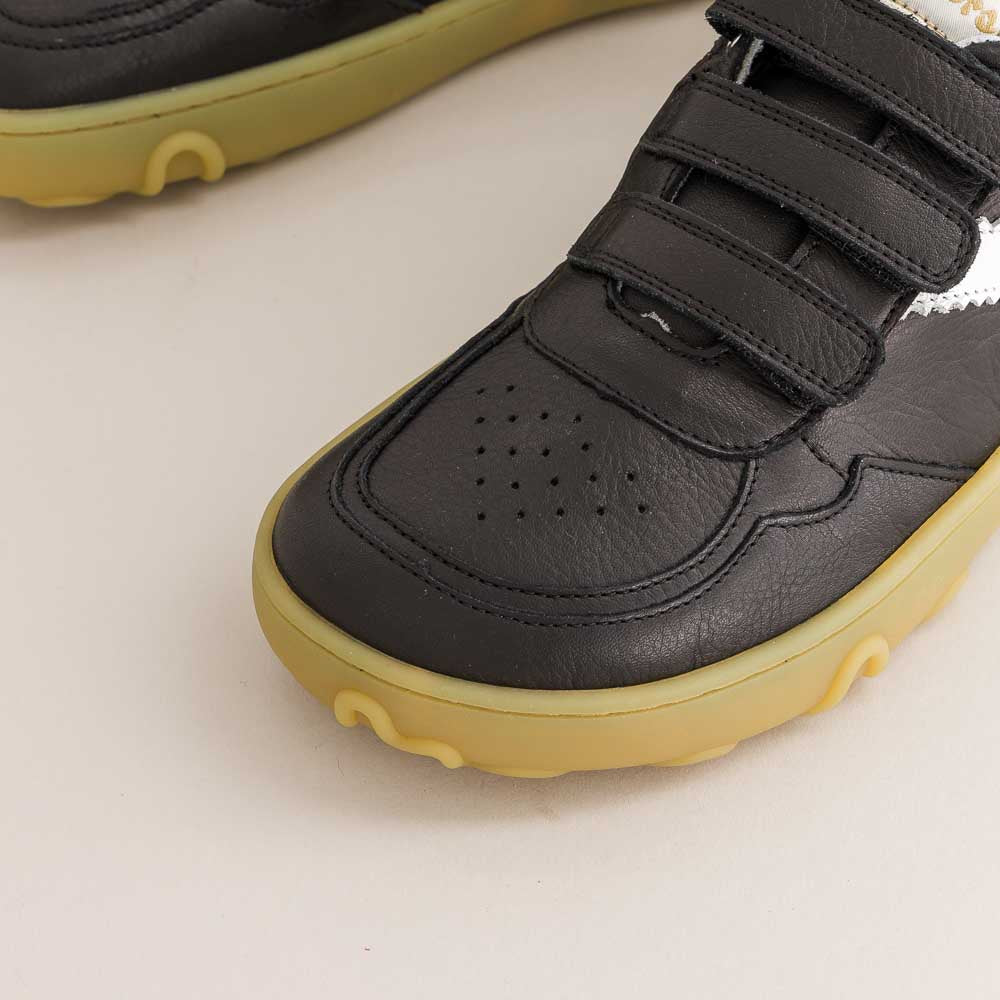 A close-up of a black children's barefoot sneaker with three hook-and-loop straps and a flexible yellow sole, shown on a light off-white background.