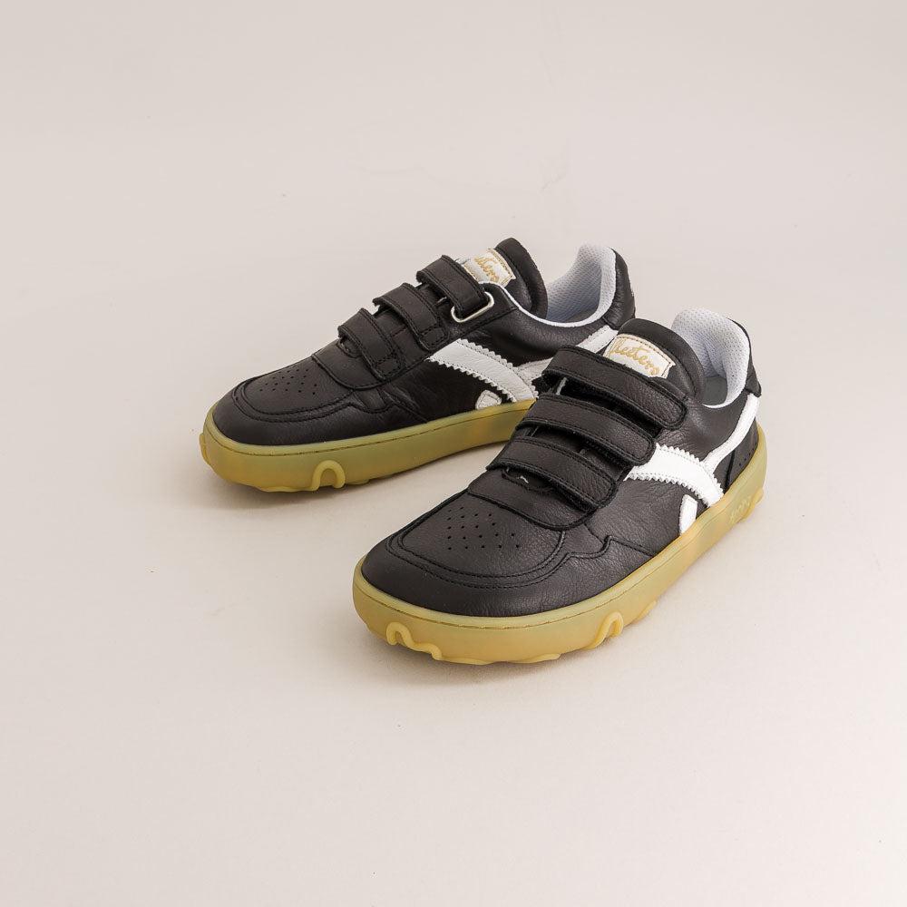 A pair of black kids' barefoot sneakers with hook-and-loop straps, white side stripes, and gum soles, photographed on a light beige studio background.
