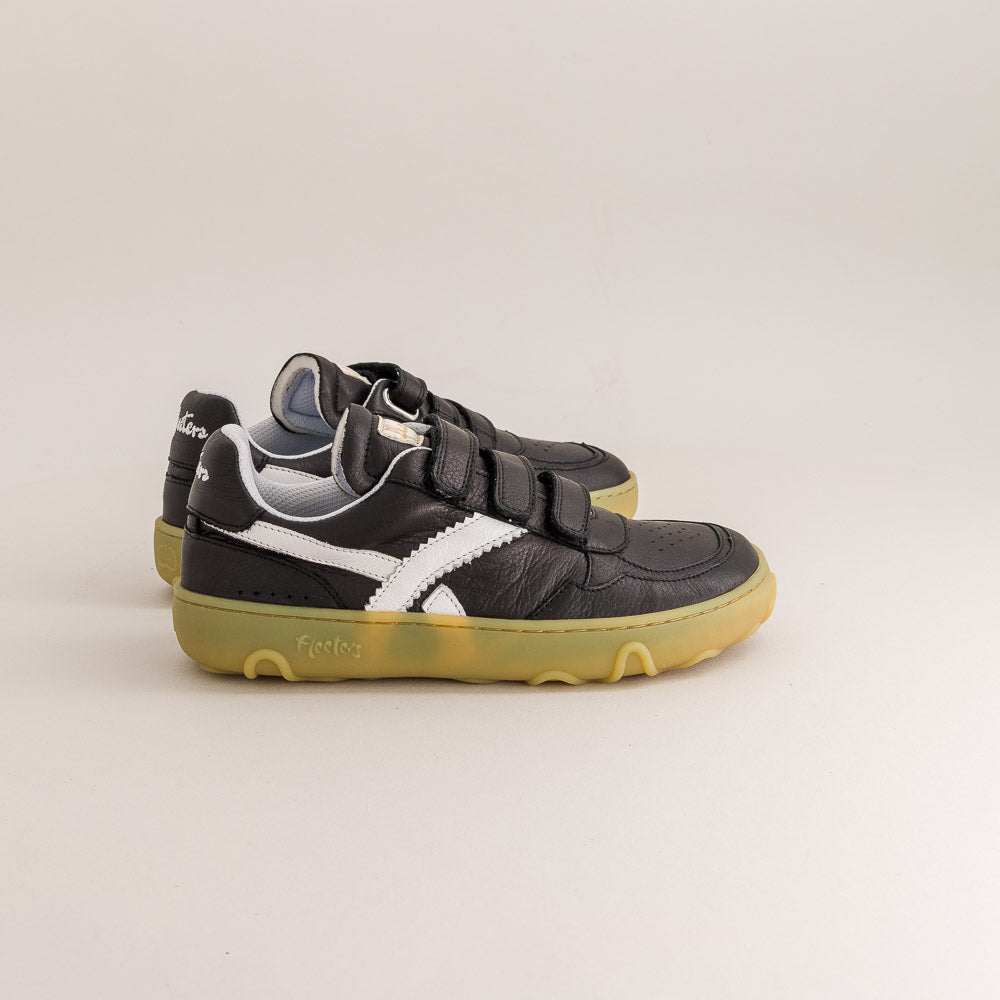 A pair of black leather barefoot sneakers with white side stripes, double hook-and-loop straps, and gum soles, photographed on a light beige studio background.