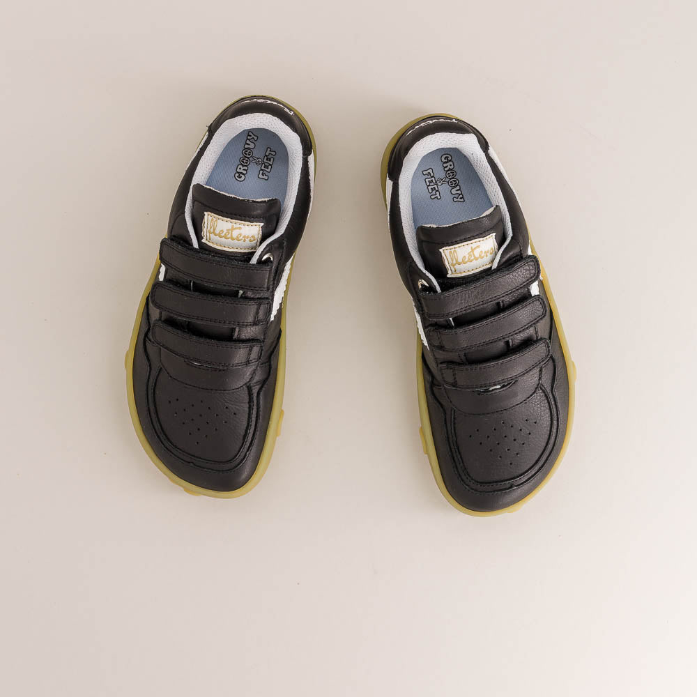 A pair of black low-top sneakers with double hook-and-loop straps, a light blue lining, and a translucent yellow sole, shown from a top-down view on a light beige background.