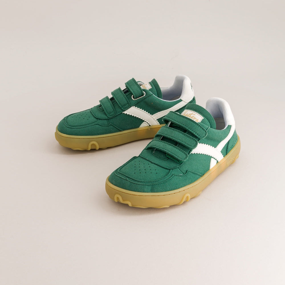 A pair of green toddler sneakers with white side stripes and yellow soles, photographed on a light beige studio background.