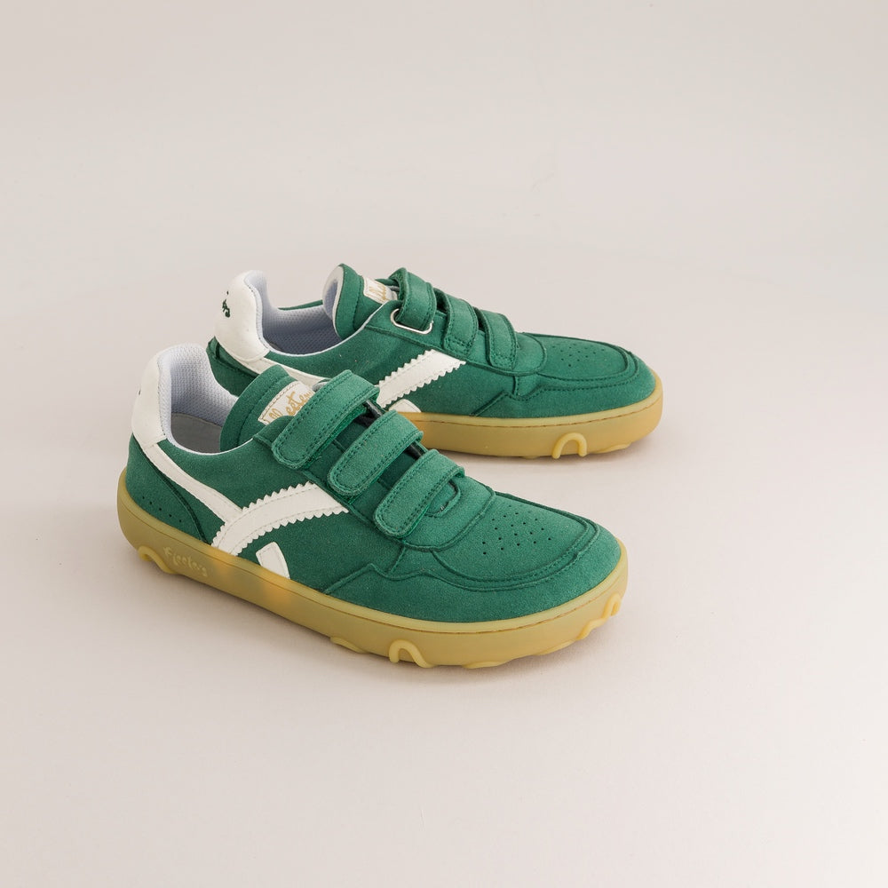 A pair of green children's sneakers with white side stripes and yellow soles, photographed on a light beige studio background.