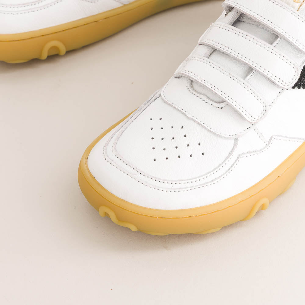 A close-up of a white low-top sneaker with hook-and-loop strap closures and a gum yellow sole, shown on a light beige background.