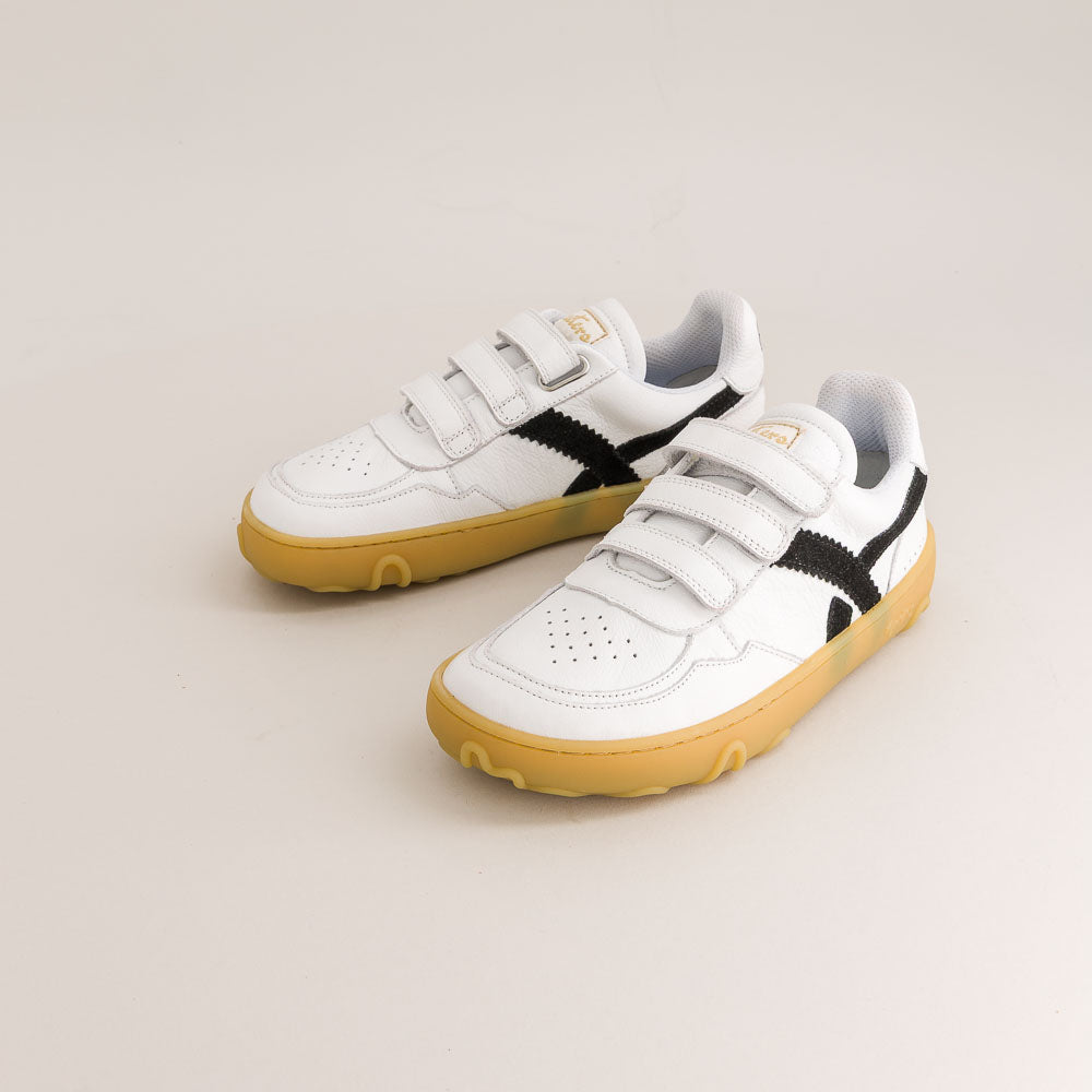 A pair of white kids' barefoot sneakers with hook-and-loop straps, black side stripes, and gum soles, photographed on a light beige studio background.