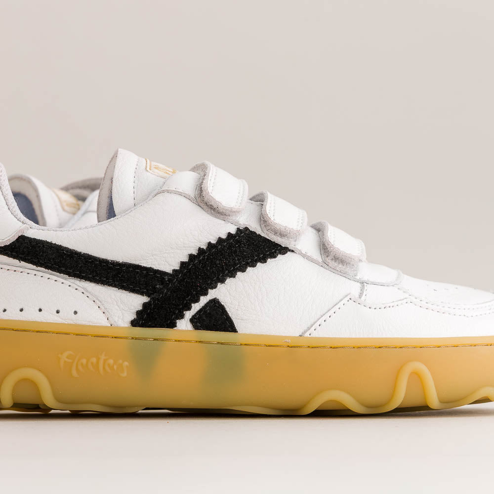 A white leather low-top sneaker with black side stripes, double hook-and-loop strap closures, and a translucent yellow gum sole.