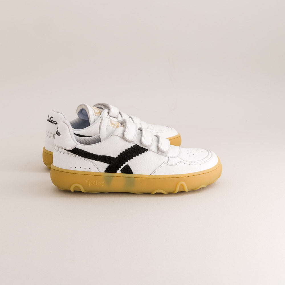 A pair of white barefoot sneakers with double hook-and-loop straps, a black side stripe, and a thick yellow gum sole, photographed on a light beige studio background.