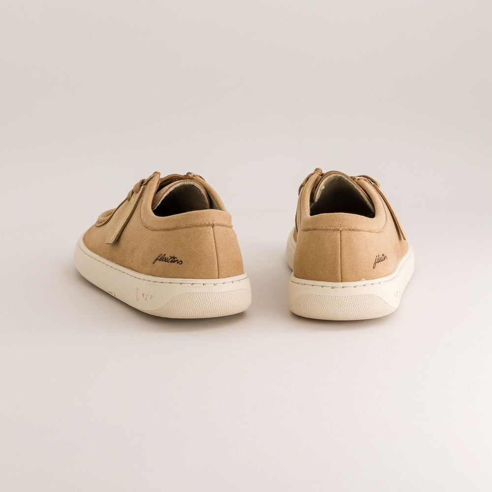 A pair of tan suede low-top sneakers shown from the back on a light gray studio background. The shoes feature tan laces, a tan heel tab with a small script logo, and off-white soles.