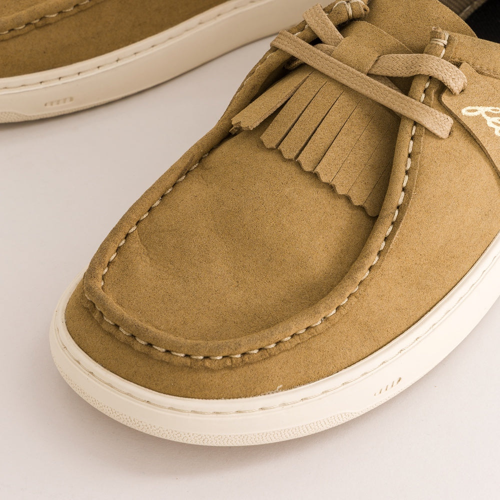 A close-up of a tan suede moccasin-style loafer with a fringe detail across the top, matching tan laces, and a thick white sole, photographed on a clean white background.