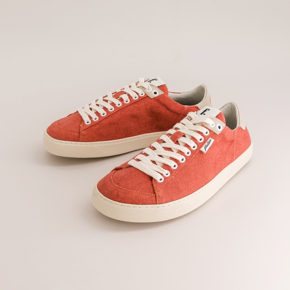 A pair of low-top canvas barefoot sneakers in a warm coral-red color, featuring off-white laces and thick off-white rubber soles, photographed on a light beige studio background.