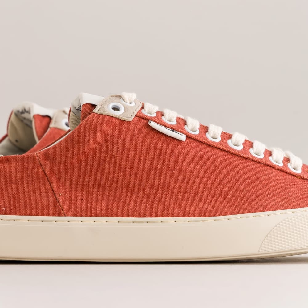 A close-up side view of a low-top canvas barefoot sneaker in a muted terracotta red, featuring white laces, white eyelets, and a thick cream-colored rubber sole.