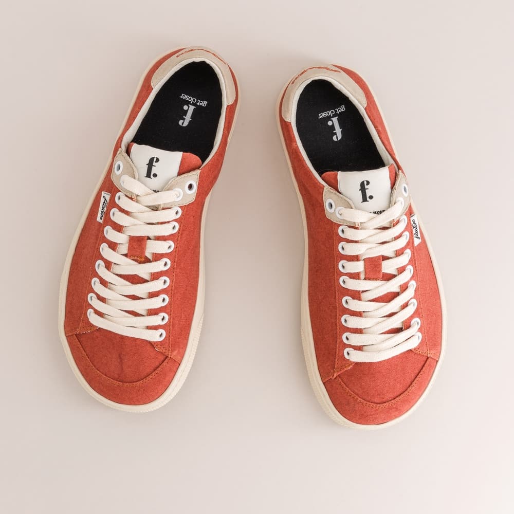 A pair of low-top canvas barefoot sneakers in a muted terracotta red, featuring white laces, white rubber soles, and a white tongue tab with a black logo. The shoes are photographed from a top-down perspective on a light beige background.