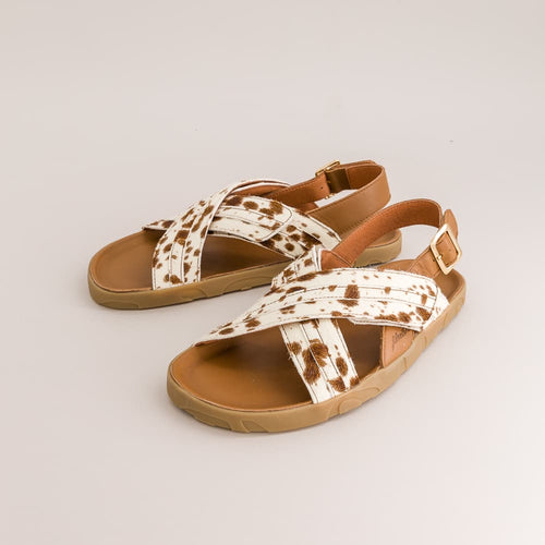 A pair of cow-print leather sandals with brown straps and tan soles, displayed on a light gray background.