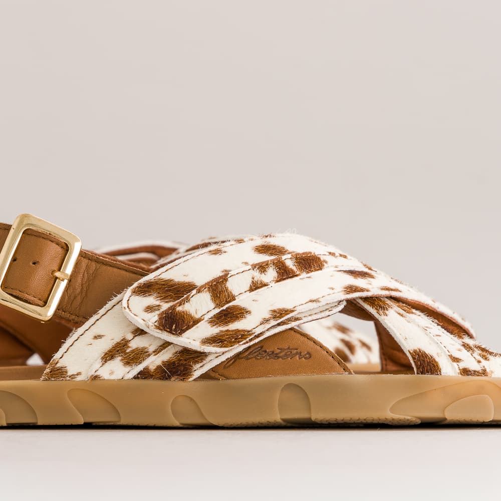 A close-up side view of a single sandal featuring brown and white cow-print straps, a tan leather buckle strap, and a tan sole, set against a neutral light gray background.