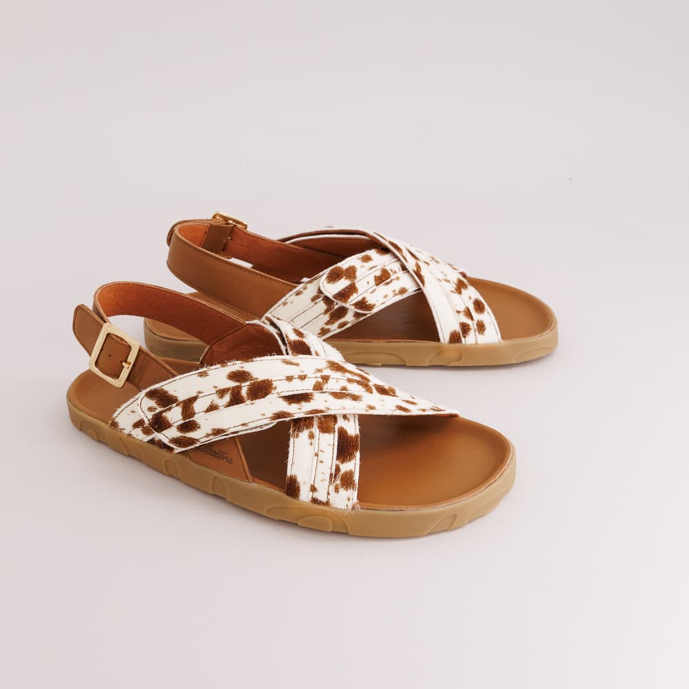 A pair of women's flat sandals with brown and white cow-print straps, tan footbeds, and light brown soles, shown on a light gray background.