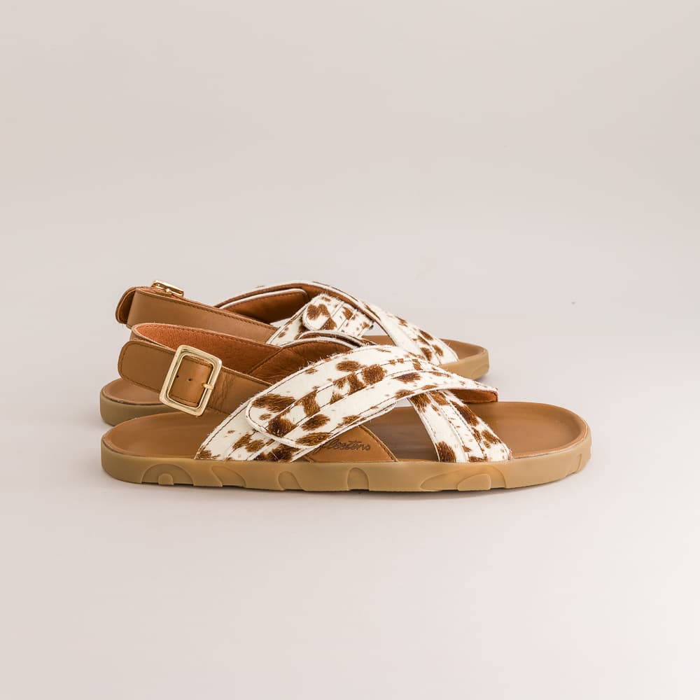 A pair of women's strappy sandals with brown and white cow-print straps, tan soles, and gold-tone buckle details, shown against a light gray studio background.