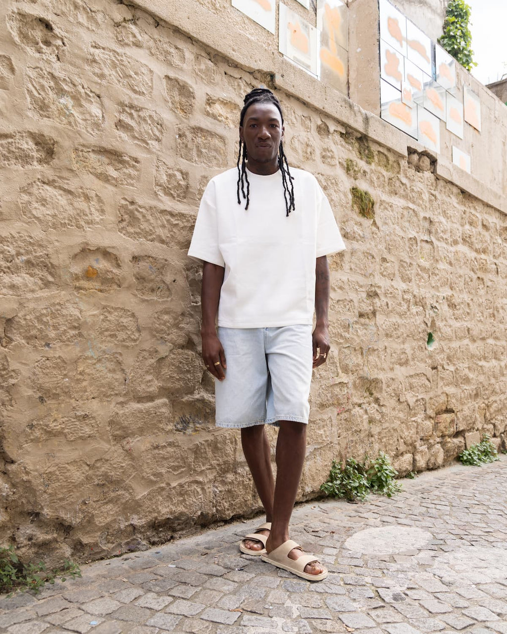 Person wearing a white t-shirt and light shorts standing against a stone wall.