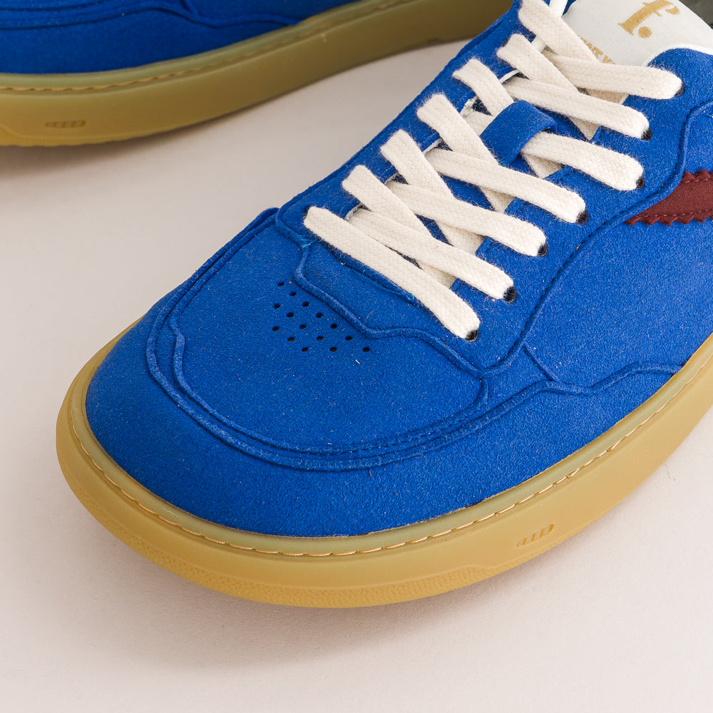 A close-up of a blue suede sneaker with white laces, a tan gum sole, and a small red side stripe, photographed on a light neutral background.