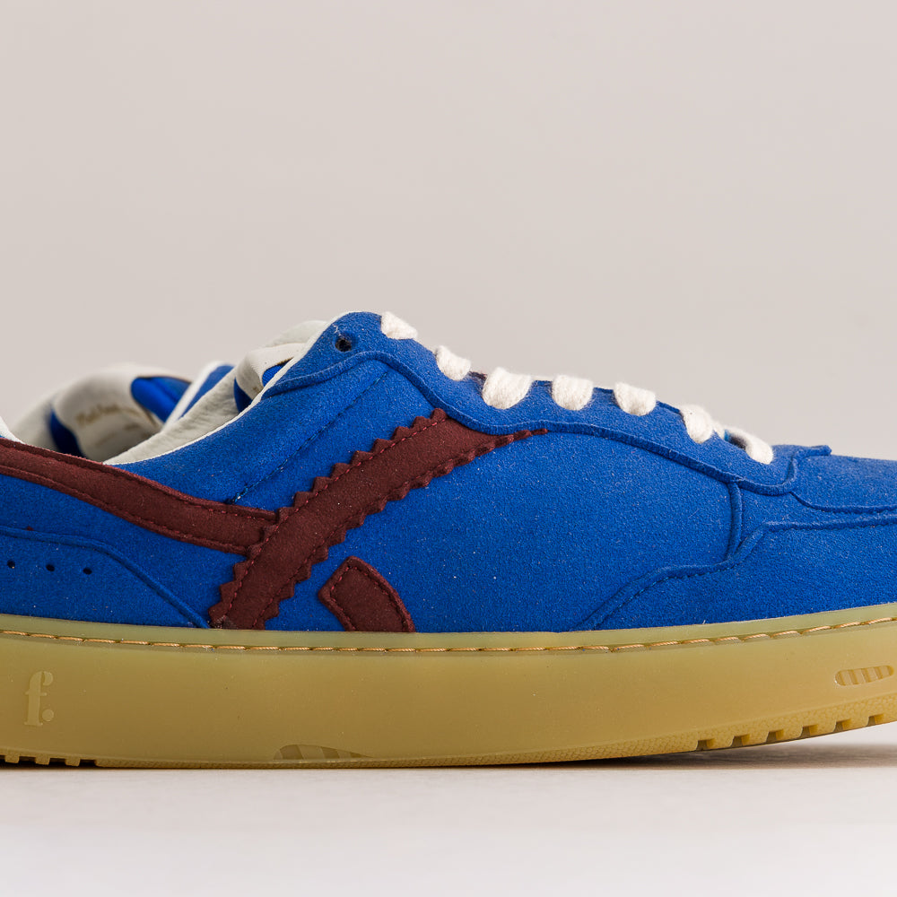A low-top sneaker in royal blue suede with dark brown side stripes, white laces, and a thick gum-colored sole, shown in a side profile against a light gray background.