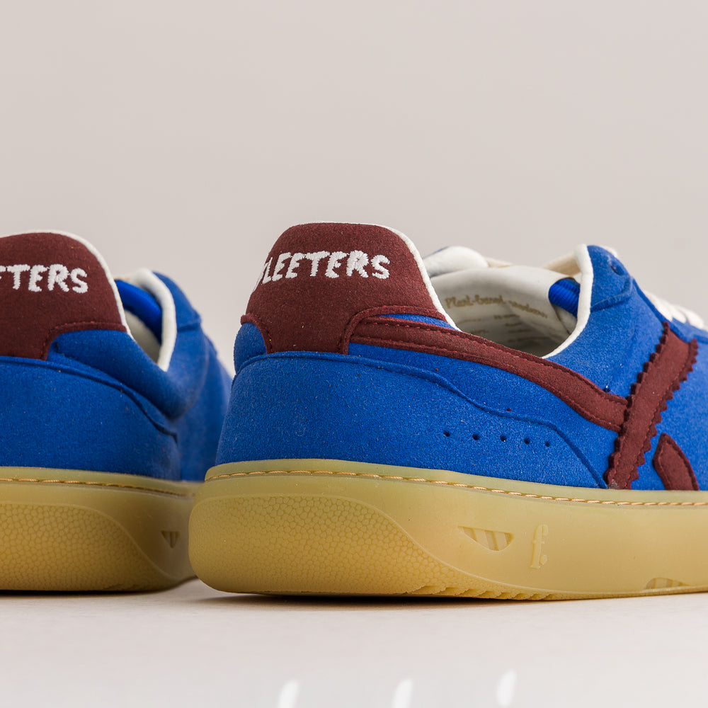 Blue suede and canvas sneakers with burgundy heel tabs and a gum sole, shown from the side and back on a light beige background.