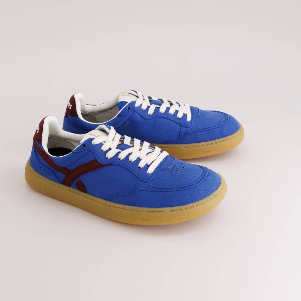 A pair of blue suede barefoot sneakers with white laces, a dark red side stripe, and gum soles, photographed on a light gray studio background.
