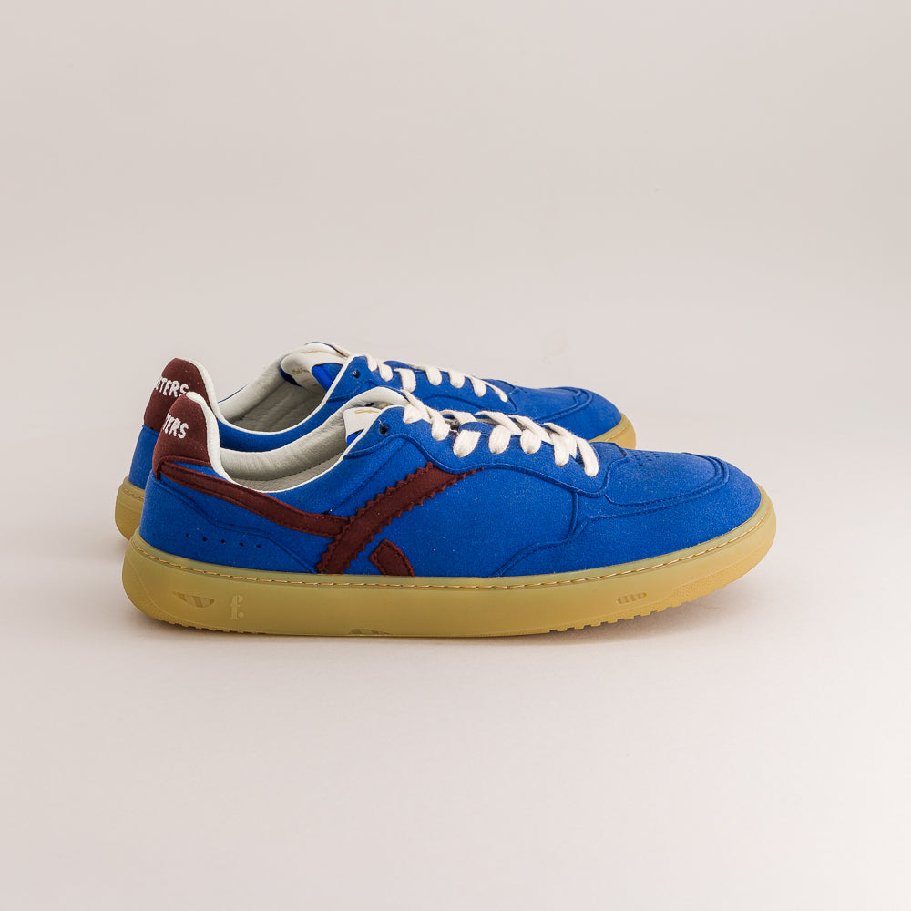 A pair of blue low-top sneakers with white laces, a brown side stripe, and a gum sole, photographed on a light beige studio background.