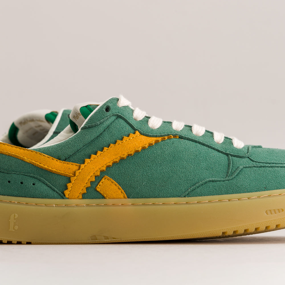 A green suede low-top sneaker with a bright yellow zigzag side stripe, white laces, and a gum-colored sole, shown in a side profile against a light gray background.