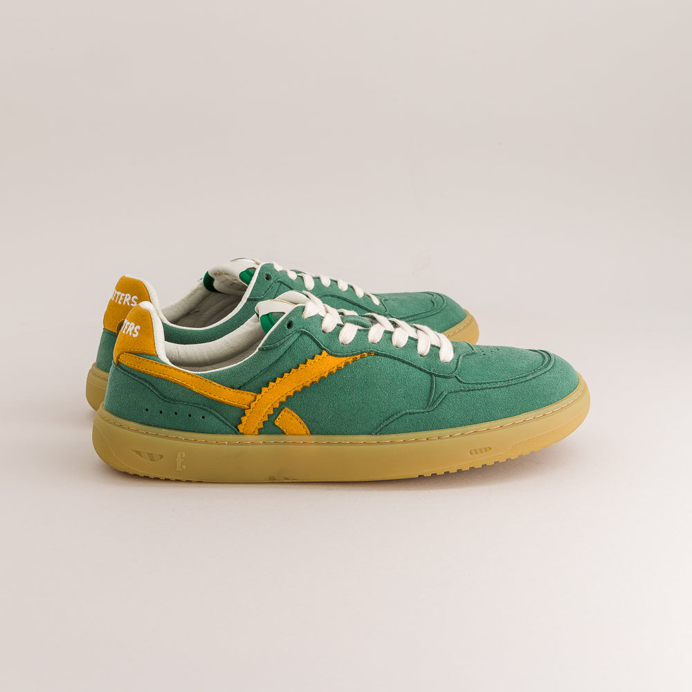 A pair of green suede sneakers with yellow side stripes, white laces, and a gum sole, photographed on a light beige studio background.
