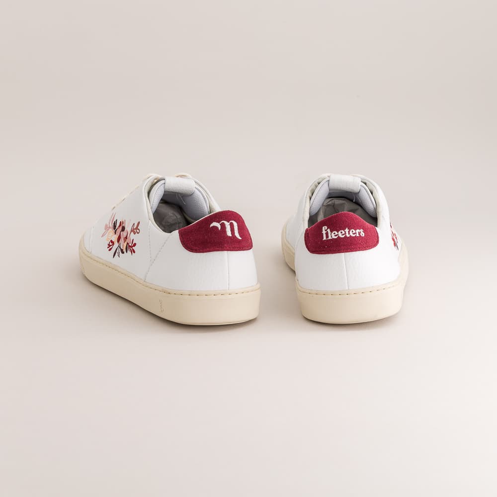 A pair of white leather barefoot sneakers with cream soles, featuring a floral embroidery on the side in pink, red, and green, and a burgundy heel tab with white lettering.
