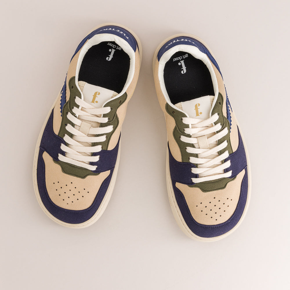 A pair of low-top barefoot sneakers shown from a top-down view on a light beige background. The shoes feature a cream-colored leather base, navy blue suede overlays, olive green panels, and white laces.