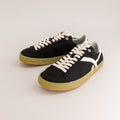 A pair of black canvas low-top sneakers with off-white laces, a white side stripe, and a gum-colored sole, photographed on a clean light beige studio background.