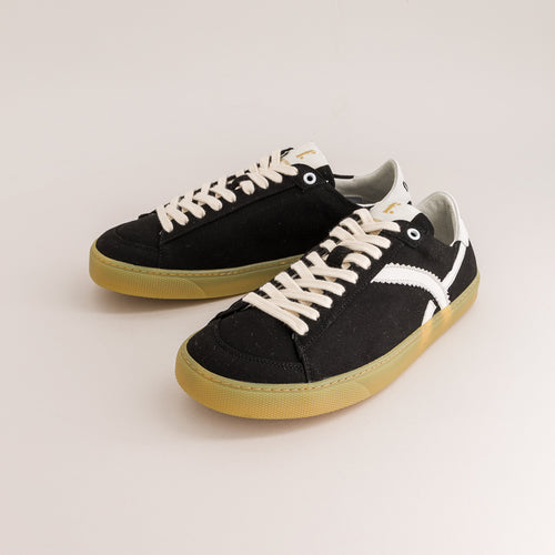 A pair of black canvas low-top sneakers with off-white laces, a white side stripe, and a gum-colored sole, photographed on a clean light beige studio background.