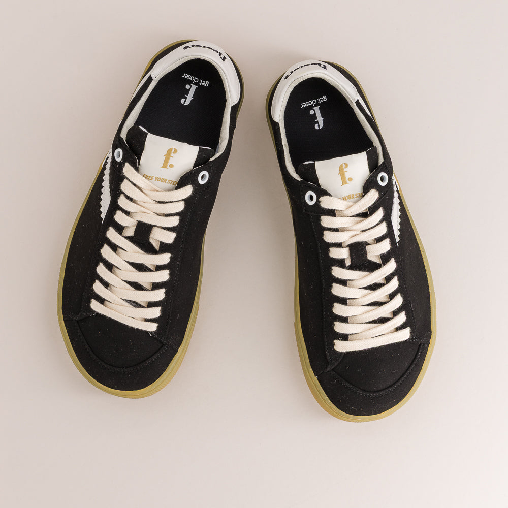 A pair of black low-top sneakers with off-white laces, white side stripes, and gum soles, photographed from a top-down angle on a light beige background.