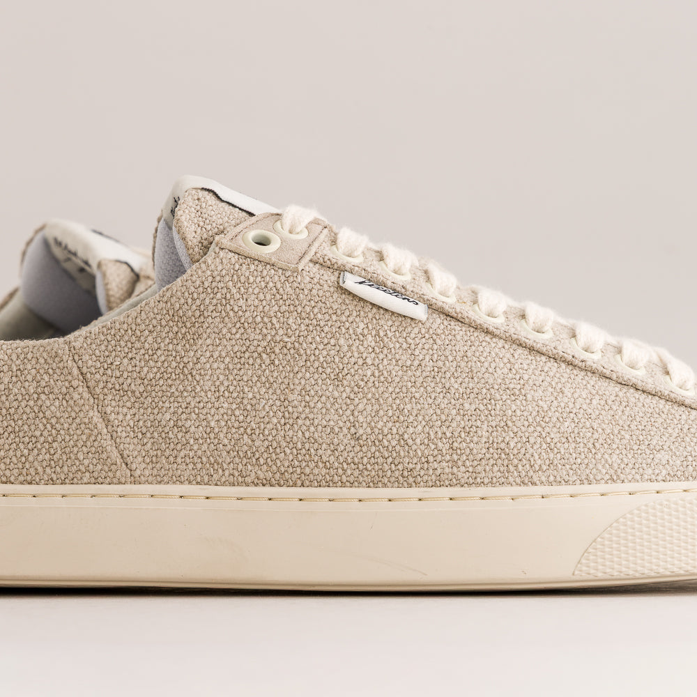 A close-up side view of a low-top wool sneaker in a light beige color, featuring matching beige laces and a cream-colored rubber sole, set against a neutral light gray background.